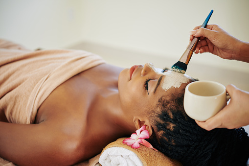 Close-up image of beautician applying deep clearing clay mask on face of Black young woman
