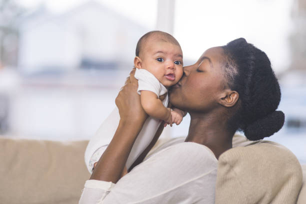 Beautiful African American mother holds newborn baby in the living room A beautiful young African American mother gently holds her infant daughter up in the air with both hands and kisses her cheek. The baby's eyes are wide open and she looks happy. They are sitting on a couch in their living room. newborn stock pictures, royalty-free photos & images