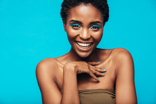 Close up of beautiful young african woman with vivid makeup smiling against blue background. Female model wearing artistic makeup looking at camera.
