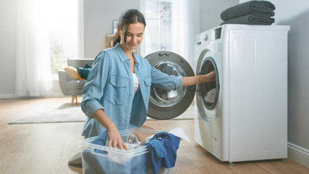 美しく、幸せなブルネットの若い女性は家庭的なジーンズの服で洗濯機に向かって来る。彼女は汚れた洗濯物で洗濯機をロードします。モダンなインテリアが施された明るく広々としたリビ� - 洗濯機 ストックフォトと画像