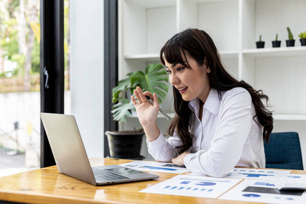 Beautiful Asian businesswoman sitting in her private office, she is talking to her partner via video call on her laptop, she is a female executive of a startup company. Concept of financial management stock photo
