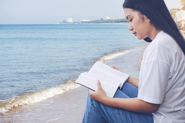 beautiful asian woman reads a book or the holy bible while sitting at the seaside. relax reading book with seascape background. - havenmeester stockfoto's en -beelden
