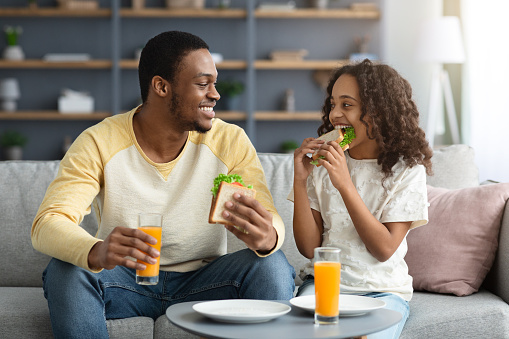 Beautiful black family father and daughter having snack at home, eating healthy fresh sanwiches and drinking orange juice, having pleasure conversation, sitting on couch in living room