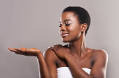 Skin hydration concept. Beautiful african american girl demonstrating something on her open palm, standing wrapped in towel over grey background