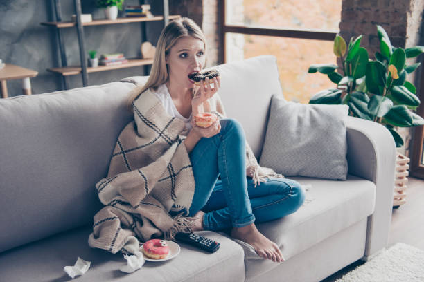 beautiful blonde woman sitting on couch in living room under blanket eating chokolate donat watching something exciting interesting on television having health problem - come e sente imagens e fotografias de stock