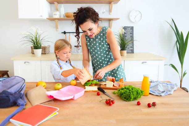 Beautiful brunette mother and her daughter packing healthy lunch and preparing school bag stock photo