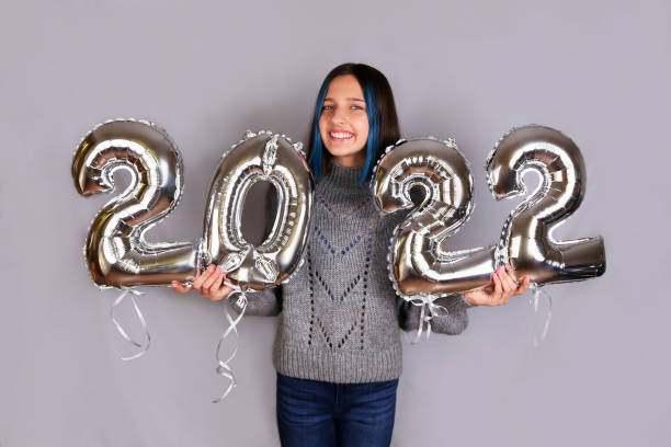 A beautiful cheerful brunette on a gray background holds silver balloons from the number 2022 in her hands. Happy A beautiful cheerful brunette on a gray background holds silver balloons from the number 2022 in her hands. Happy New Year 2022. new years eve girl stock pictures, royalty-free photos & images