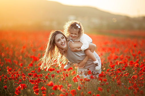 Beautiful couple mother and cute daughter are walking in field of red poppy flowers over yellow sunset lights