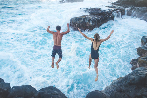 Beautiful couple jumping off cliff into ocean Beautiful couple jumping off a cliff into the turbulent ocean cliff jumping stock pictures, royalty-free photos & images