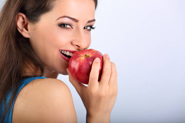 Beautiful excited makeup brunette woman biting the red tasty apple and looking happy with empty space blue background. Closeup portrait stock photo