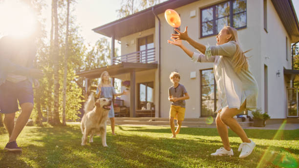 4つのプレイの美しい家族は裏庭の芝生にハッピーゴールデンレトリバー犬とおもちゃのボールをキャッチ。牧歌的な家族は、夏の家の裏庭で忠実な血統犬の屋外で楽しんでいます。 - 庭 ストックフォトと画像