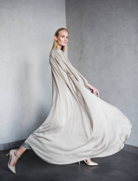 Beautiful female fashion model wearing modern loose dress. Gorgeous young blonde woman walking and posing in a studio while wearing a luxury abaya made of a long grey fabric. high fashion model stock pictures, royalty-free photos & images
