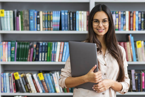 mooie vrouwelijke student met laptop die op de achtergrond van de bibliotheekboekenplanken glimlacht het leren en onderwijs online concept - academy stockfoto's en -beelden