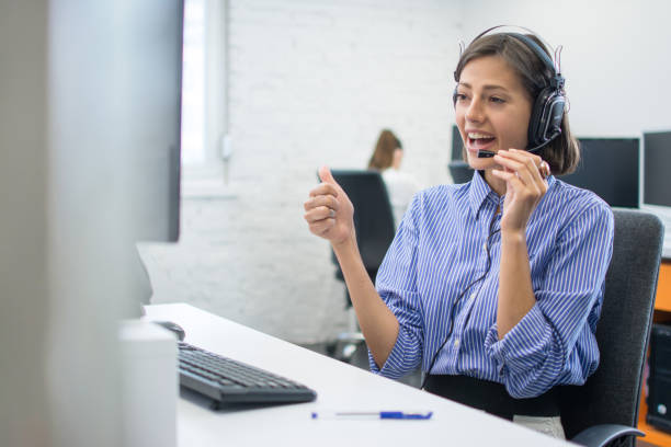 Beautiful friendly female costumer service operator showing thumb up to computer screen during video call in call centre stock photo