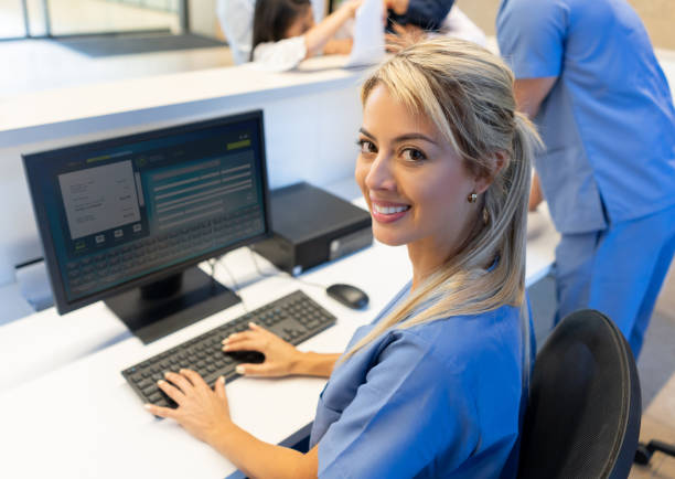 vacker vänlig sjuk sköterska i receptionen på sjukhuset arbetar på datorn medan inför kameran leende - vårdklinik bildbanksfoton och bilder