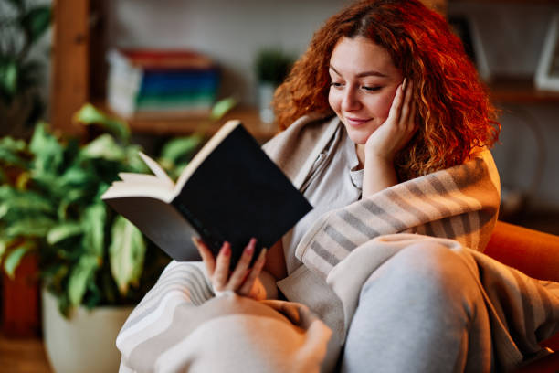 A beautiful ginger girl sitting in the living room wrapped in a blanket and reading an interesting book in the morning. stock photo
