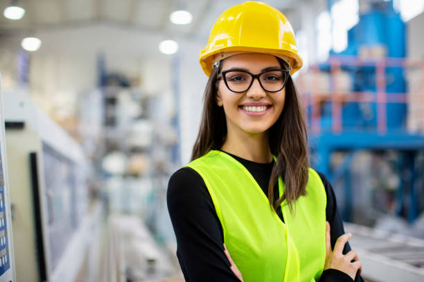 Beautiful girl at factory in a vest and helmet smiling. stock photo