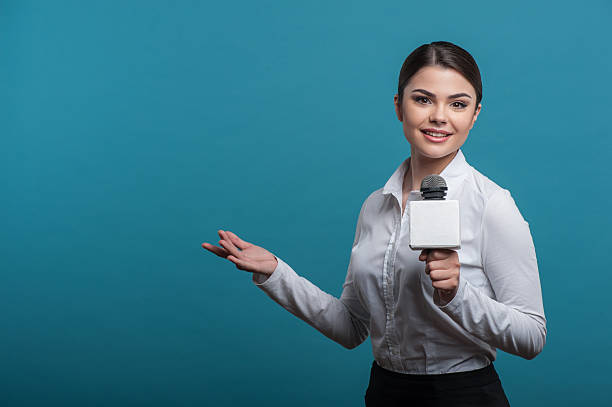 Beautiful girl TV journalist with pretty smile is reporting Waist up portrait of elegant woman reporter with brown hair, who interviews and is smiling and looking at the camera holding the microphone and making a gesture with her right hand, isolated on a blue background and there is copy place in the left side journalist stock pictures, royalty-free photos & images