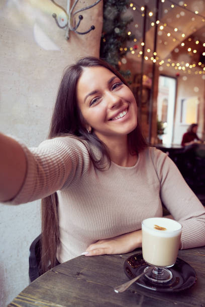 Beautiful happy girl taking a selfie in cafe during Christmas holidays, smiling and looking at phone. Brunette woman with long hair drinks cappuccino coffee, latte stock photo