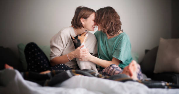 Beautiful happy lesbian couple in pajamas sitting on bed in the morning with pet dog,  tenderly kissing and hugging stock photo