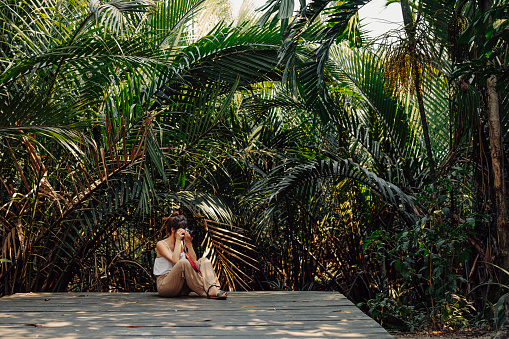 Beautiful young Caucasian woman sitting on a wooden pathway while enjoying the tropical atmosphere surrounded by palm trees in a tropical public park She is taking a photo with her camera