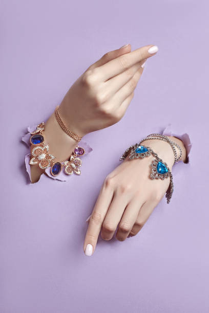 Beautiful jewelry on the hands of women, hands with bracelets and necklaces are sticking out from torn violet purple paper background. Advertising jewelry and jewelry, copy space Beautiful jewelry on the hands of women, hands with bracelets and necklaces are sticking out from torn violet purple paper background. Advertising jewelry and jewelry, copy space gold ring on finger stock pictures, royalty-free photos & images