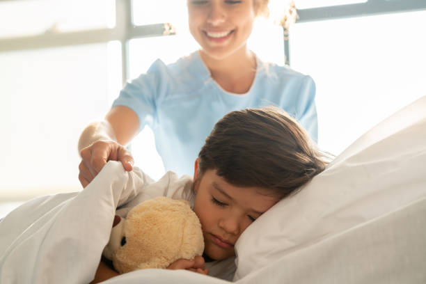 hermoso niño durmiendo con su oso de peluche en una cama de hospital mientras que la enfermera lo cubre correctamente con cubiertas sonriendo - teddy ray fotografías e imágenes de stock
