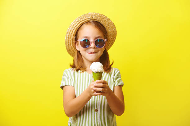 güzel küçük kız bir dondurma tutan ve onu yemek için gidiyor, dudaklarını yalama, bir yazlık elbise giymiş, hasır şapka ve güneş gözlüğü, sarı izole ayakta. - ice cream stok fotoğraflar ve resimler