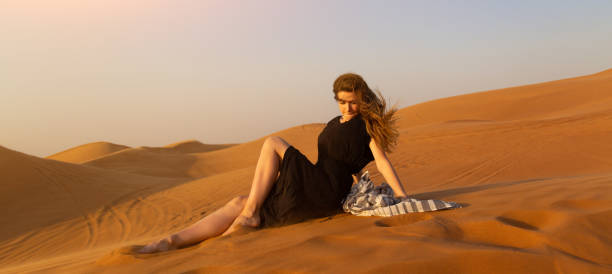 Beautiful lonely girl at sunset in the desert. A young woman in a black dress sits on the sand in the desert meeting the sunset hot arabian women stock pictures, royalty-free photos & images