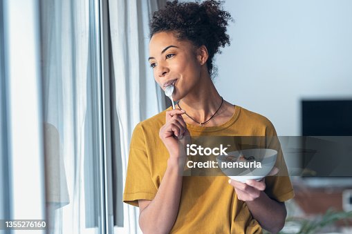 istock Beautiful mature woman eating cereals and fruits while standing next to the window at home. 1355276160