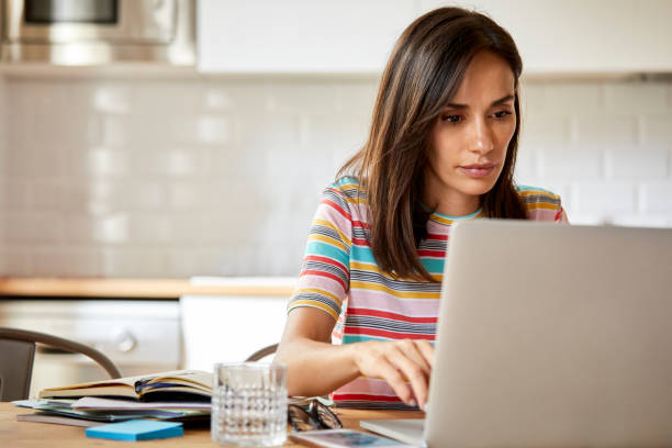 Close-up of beautiful woman using laptop while sitting at table. Confident mid adult female with long brown hair is in domestic kitchen. She is in striped t-shirt at home.