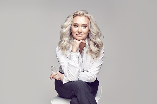 A pretty older woman with beautiful and healthy blonde hair and clear skin, looks beautiful, dressed in a strict and stylish business image, looks at the camera subtly, sits on a chair on a grey isolated background.