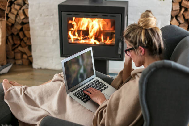 Beautiful middle-aged woman next to the fireplace relaxes in the living room and works on laptop from house stock photo