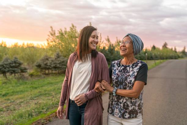 Beautiful mixed race mother and daughter relaxing outdoors together A senior woman with cancer walks with her adult daughter at sunset down a rural road. They are relaxing and staying active together. The affectionate pair are talking and walking with arms linked. cancer illness stock pictures, royalty-free photos & images