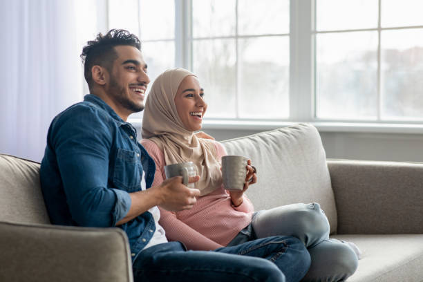 Beautiful muslim couple drinking tea, looking at copy space Beautiful muslim couple sitting on sofa at home, drinking tea, looking at copy space. Happy young middle-eastern family wife and husband drinking coffee and watching TV, enjoying time together hot middle eastern women stock pictures, royalty-free photos & images