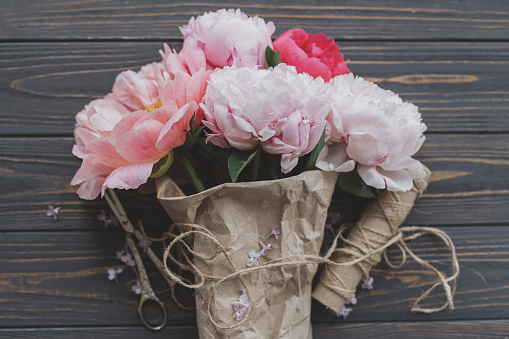 Beautiful stylish peony bouquet in paper, twine and scissors on rustic dark wooden background, top view. Happy Mothers day greetings. Pink and white peonies flowers and lilac petals