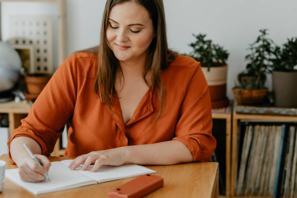 Beautiful  plus size Woman Sitting in her Living Room and Writing a Diary A smiling plus size woman sitting at her desk and writing a journal. writing activity stock pictures, royalty-free photos & images