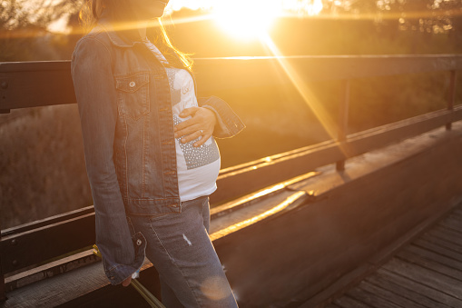 Beautiful pregnant woman standing on footbridge in the park and enjoys the sunset.. She wears jeans and caresses her stomach with her hands