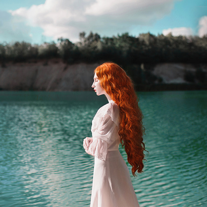 Beautiful red-haired woman in a luxurious renaissance dress on the background of a lake with blue water