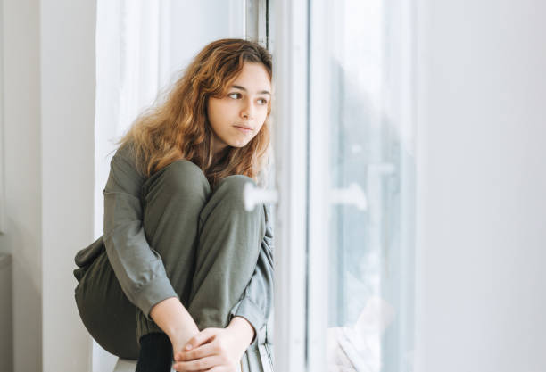 Beautiful sad unhappy teenager girl with curly hair sitting on the window sill stock photo