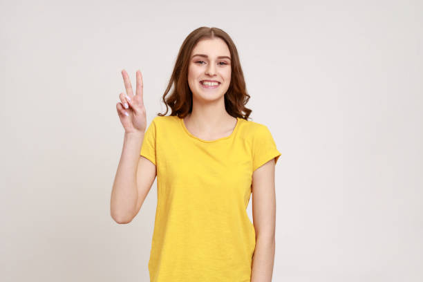 Beautiful satisfied teenager girl showing v sign symbol of peace with fingers, looking at camera with happy smile, celebrating triumph. stock photo