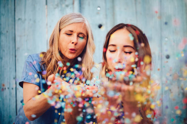 Cheerful grandmother celebrating with teenage granddaughter by blowing multi-colored confetti in the city