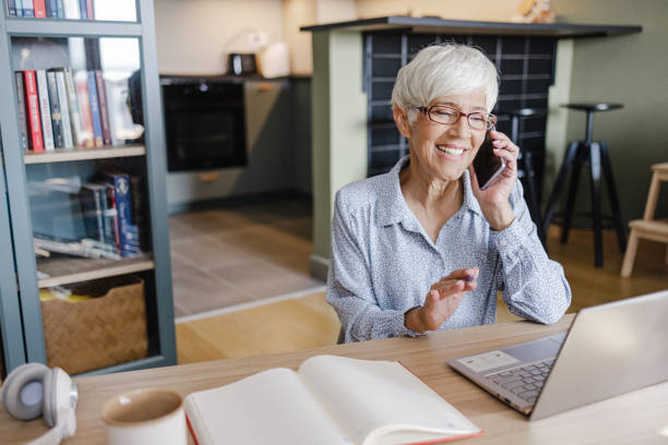 A beautiful senior woman is talking on the phone in the living room while working on the laptop stock photo