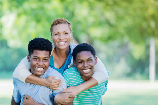 Beautiful single mom smiles for camera with two teenage sons A beautiful African American mom stands behind her two teenage sons in embrace as they all smile for the camera outdoors. mother and teenage son stock pictures, royalty-free photos & images