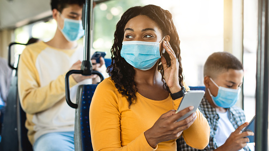 Portrait of happy smiling black female in medical face mask traveling on public transit, listening to music touching wireless earphones and using cellphone, looking away at window, sitting inside