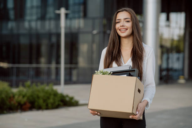 Beautiful smiling brunette girl in a shirt stands in front of the glass modern building where she worked in hands holding a box with packed things, promotion to a position, the joy of quitting Beautiful smiling brunette girl in a shirt stands in front of the glass modern building where she worked in hands holding a box with packed things, promotion to a position, the joy of quitting quitting a job photos stock pictures, royalty-free photos & images
