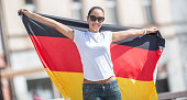 Beautiful smiling female fan in sunglasses holds a German flag behind her outdoors.