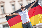 Beautiful smiling girl in sunglasses holds a German flag behind her outdoors.