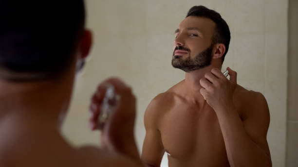 Beautiful smiling man spraying perfume on body bathroom, preparing for romantic date stock photo