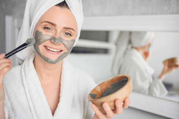 Beautiful smiling woman at home in bathrobe with a towel applying face clay mask against acne to rejuvenate problem skin stock photo